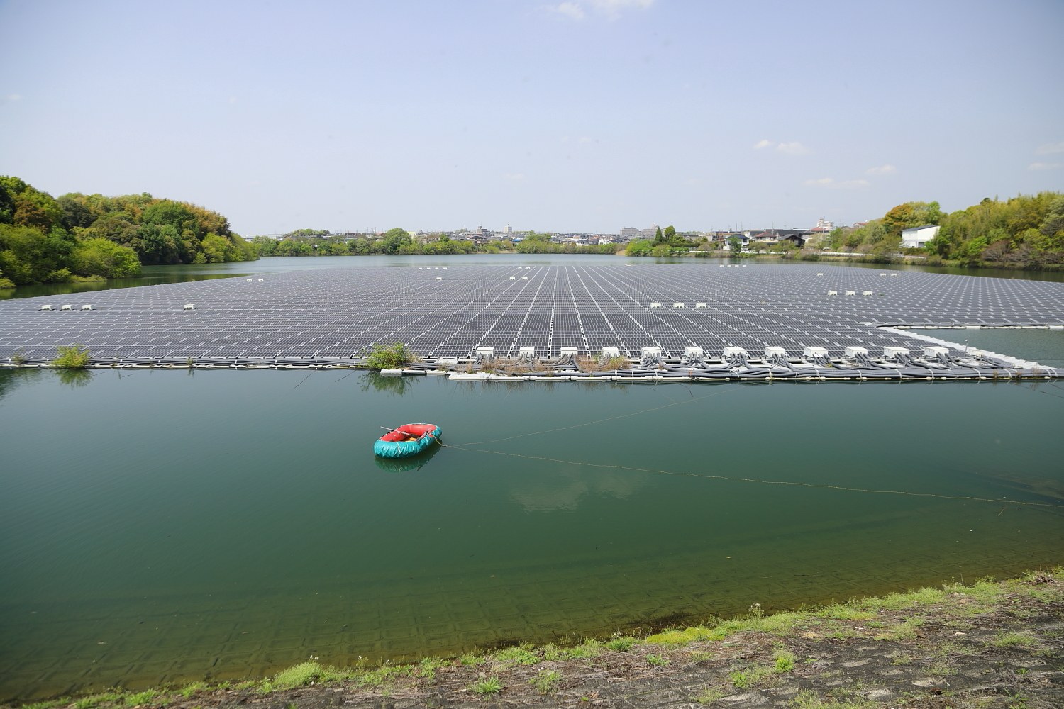 Kleines Boot mit Solarpanelen auf der Oberfläche, das auf Wasser schwimmt und von Grün umgeben ist, mit Gebäuden und einem klaren blauen Himmel im Hintergrund.
