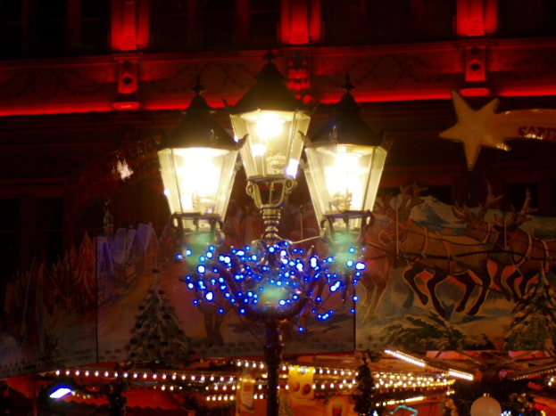 Eine Stra├čenlaterne in einer Stadt bei Nacht, geschm├╝ckt mit Lichtern und Schmuck, steht vor einem Geb├Ąude und verbreitet eine festliche Atmosph├Ąre.