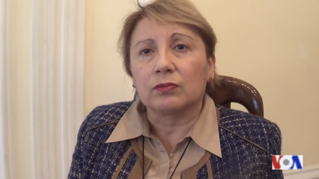 Eine Frau sitzt auf einem Stuhl vor einem Fenster und sieht besorgt aus, mit einer Wand im Hintergrund und einem Wasserzeichen im rechten unteren Eck.