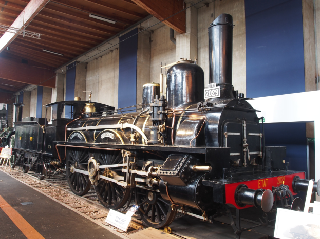 Alte Dampflokomotive in einem Museums Setting mit umgebenden Eisenbahnschienen, Informationsschildern und anderen Ausstellungen gegen eine Wand mit ├ťberkopfbeleuchtung.