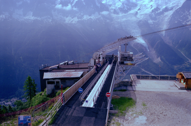 Seilbahn fährt einen Berg mit schneebedeckten Gipfeln im Hintergrund hoch, über einer Straße mit Schildern, Geländern, Vegetation, Häusern und Menschen.