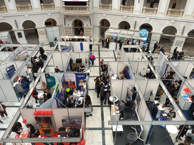 Eine große Gruppe von Menschen auf einer internationalen Messe mit Ständen, Bannern, Tischen, Stühlen und Luftballons in einem Raum mit architectural arches und einem Bildschirm im Hintergrund.