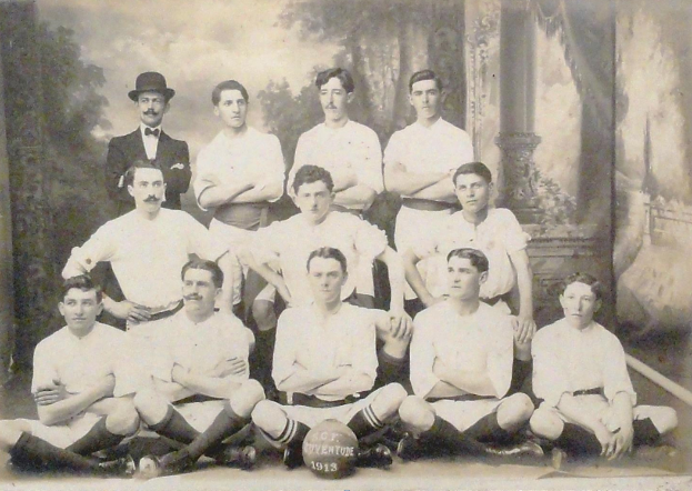 Ein Schwarz-Weiß-Foto einer Fußballmannschaft, mit einigen Spielern, die sitzen, und einigen, die stehen, einem Ball in der Mitte und Bäumen und einem klaren Himmel im Hintergrund, mit Text unten.