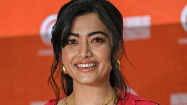 Eine Frau in einem leuchtend roten Kleid mit einem eleganten Dutt und dezentem Make-up posiert strahlend vor der Kamera, trägt schwarze High Heels und hat ein hellgelbes Banner mit der Aufschrift 'Sonam Kapoors Schönste Looks vom Cannes Film Festival' im Hintergrund.