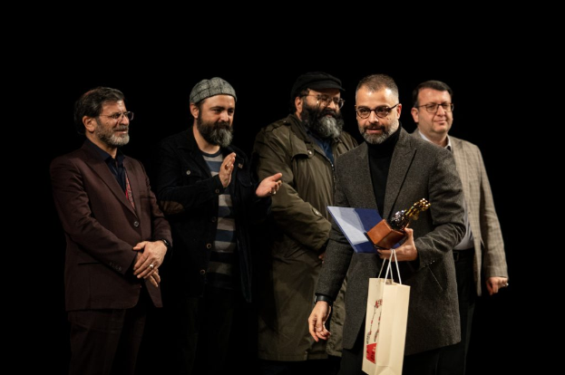 Eine Gruppe von Männern steht auf einer Bühne zusammen, einer hält eine Tasche und eine Trophäe, mit einem dunklen Hintergrund, der die Eröffnungsnacht des Berliner Filmfestivals suggeriert.