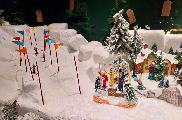 Eine festliche Weihnachtsdorfszene mit einem schneebedeckten Berg im Hintergrund, mit Bäumen, Häusern, Menschen, Fahnen, Pfählen, Lichtern, Tieren und anderen Gegenständen, die alle von Schnee bedeckt sind, unter einem dunklen, gemütlichen Ambiente.