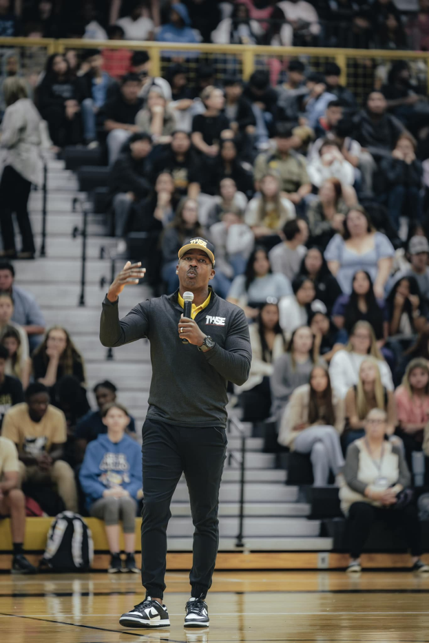 Ein Mann mit Mütze und Mikrofon steht auf einem Basketballfeld, während ein Team hinter ihm auf einer Treppe sitzt und Taschen auf dem Boden liegen.