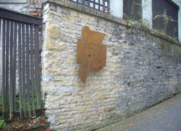 Steinmauer mit Metalltor auf der linken Seite, eine Plakette zur Einweihung des alten Rathauses und ein Gebäude mit Fenstern und Pflanzen im Hintergrund.