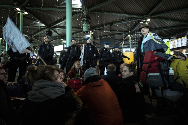 Eine Gruppe von Menschen steht vor einer Menge in einem Bahnhof, wobei einige Personen Schilder und Banner halten, was auf eine Demonstration hindeutet.