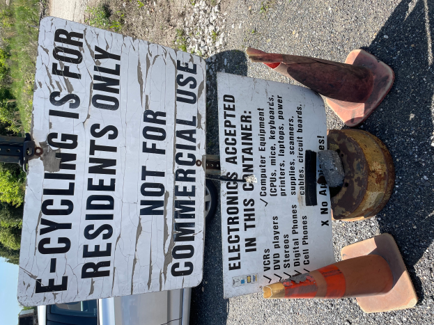 Ein Schild mit der Aufschrift "E-Bike-Fahren nur für Anwohner" steht an der Straße, umgeben von Absperrgittern und anderen Gegenständen, mit einem Fahrzeug, Bäumen und einem klaren blauen Himmel im Hintergrund.