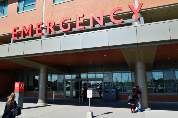 Gruppe von Menschen, die auf ein Notaufnahmegebäude mit Glaswänden, Säulen, einem Namensschild und Informationsschildern zugehen, mit einem Mülleimer in der Nähe.