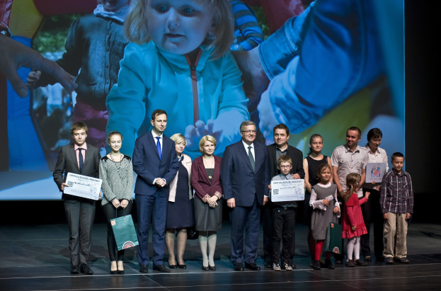 Eine Gruppe von Menschen auf einer Bühne mit Taschen und Urkunden, im Hintergrund ein Bildschirm mit dem Bild eines Kindes und anderen