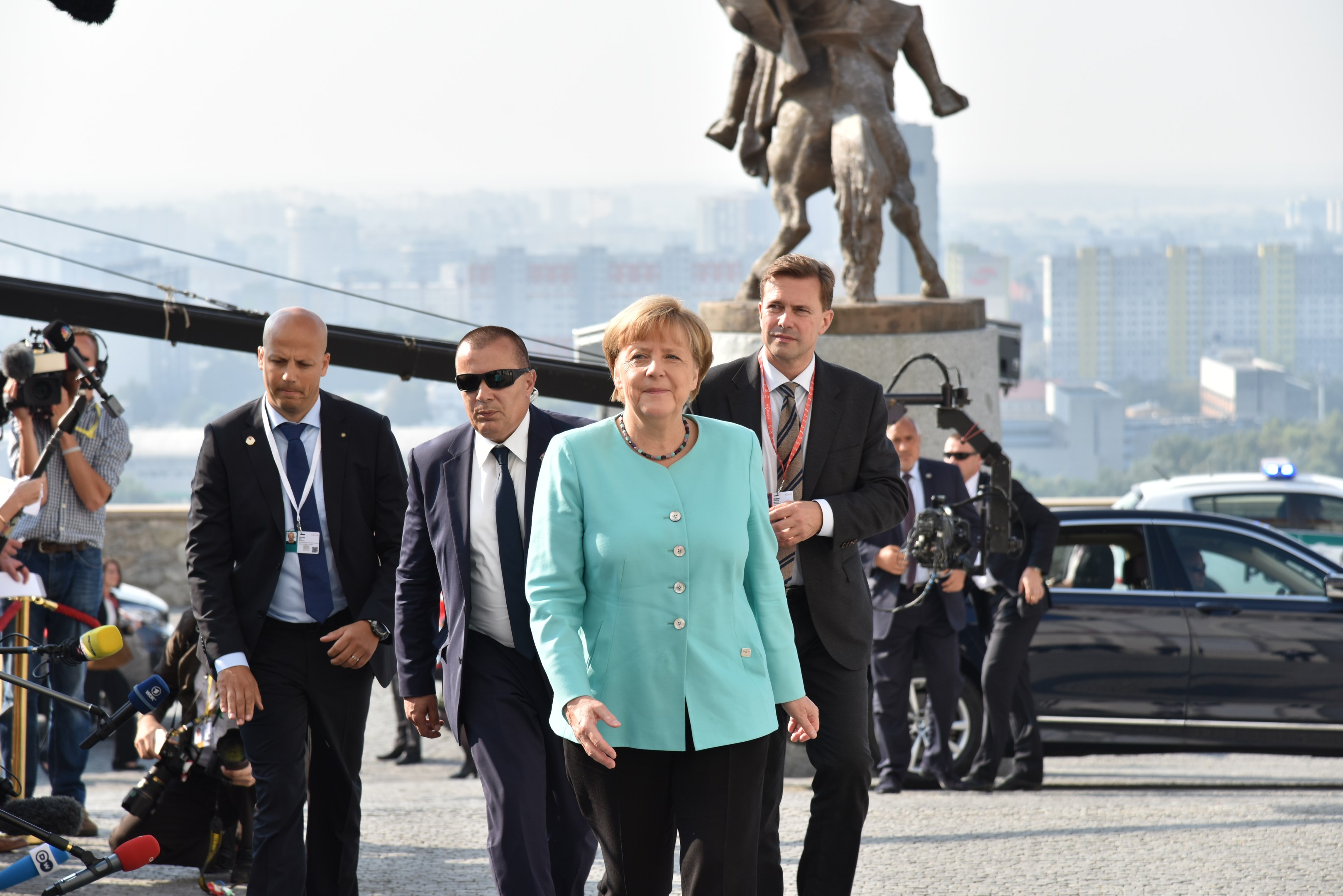 Bundeskanzlerin Angela Merkel und Bundespräsident Joachim Gauck gehen in Berlin, umgeben von Medienfahrzeugen, einer Statue, Gebäuden, Bäumen und einem klaren Himmel.