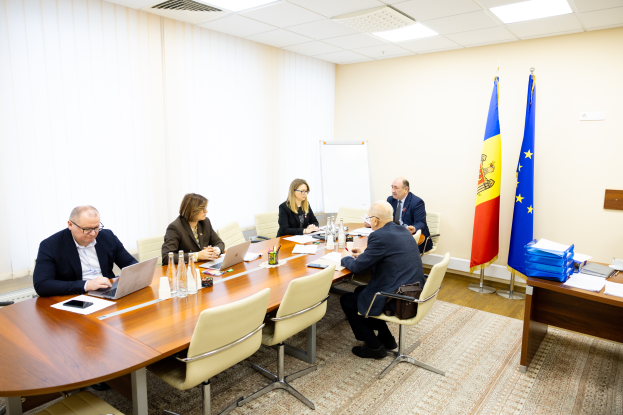 Eine Gruppe von Menschen sitzt um einen Konferenztisch mit Laptops, Flaschen, Gläsern, Papieren, Büchern und anderen Gegenständen und ist in eine Diskussion vertieft. Im Hintergrund sind Flaggen mit Stangen, eine Whiteboard an der Wand und Deckenlampen zu sehen.