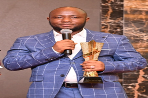 Ein Mann in einem blauen Anzug mit Mikrofon und Trophäe, der selbstbewusst in die Kamera lächelt