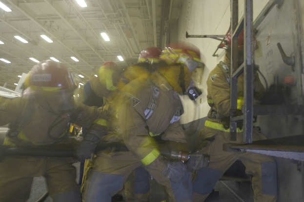 Feuerwehrleute in Schutzausrüstung arbeiten an einem Flugzeug mit einer Leiter auf der rechten Seite, umgeben von Deckenlampen.