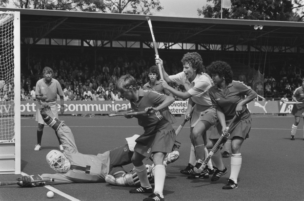 Schwarzes Bild von Frauen, die Feldhockey spielen, mit Helmen und Schlägern, ein Tor auf der linken Seite, Zuschauer auf den Tribünen, Banner, ein Schuppen, Bäume und eine Flagge im Hintergrund.