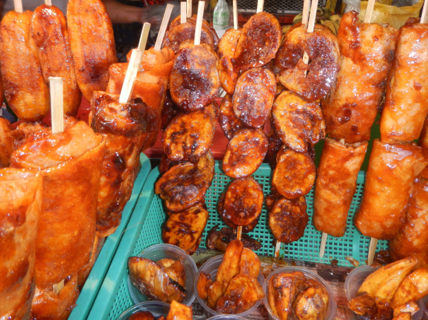 Marktstand mit Spießen von Hähnchenflügeln und verschiedenen Lebensmitteln in Plastikbehältern, umgeben von Menschen und Flaschen.