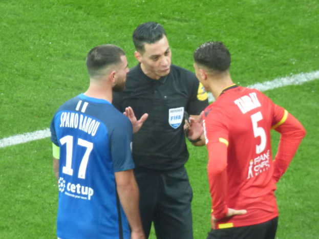 Schiedsrichter in schwarzem Hemd und Hose, der mit zwei Männern in Brille auf einem Fußballfeld spricht, einer in einem blauen und roten T-Shirt mit Text.