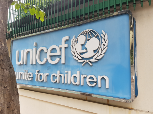 UNICEF-Logo an einer Gebäudewand mit einem Baumstamm links und einer an der Wand befestigten Tafel, Bäume im Hintergrund und ein klarer blauer Himmel.