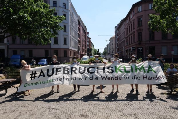 Menschen mit Masken, die ein Banner mit der Aufschrift 'Aufbruchsklima' bei einer Klimaprotest in Berlin halten, umgeben von städtischen Elementen.