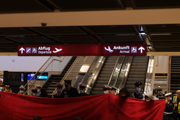 Eine Gruppe von Menschen mit Mützen und Masken steht vor einer Rolltreppe an einem Flughafen, eine rote Stoffbahn haltend und unter Schildern positioniert, wahrscheinlich beim Einsteigen in ein Flugzeug während der Coronavirus-Pandemie.