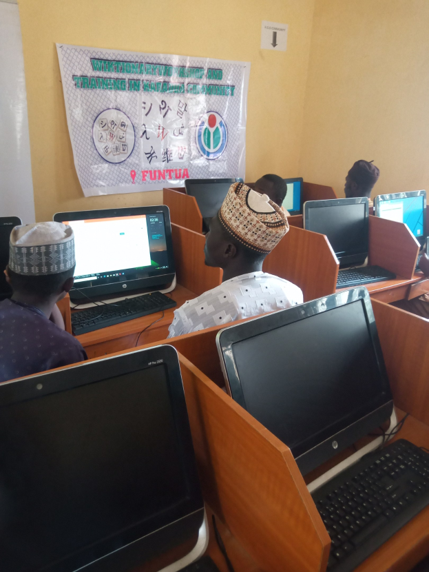 Gruppe von Menschen an Tischen mit Computerbildschirmen, Tastaturen und Mäusen bei einem Workshop zur Computertechnologie, mit einem Plakat im Hintergrund.