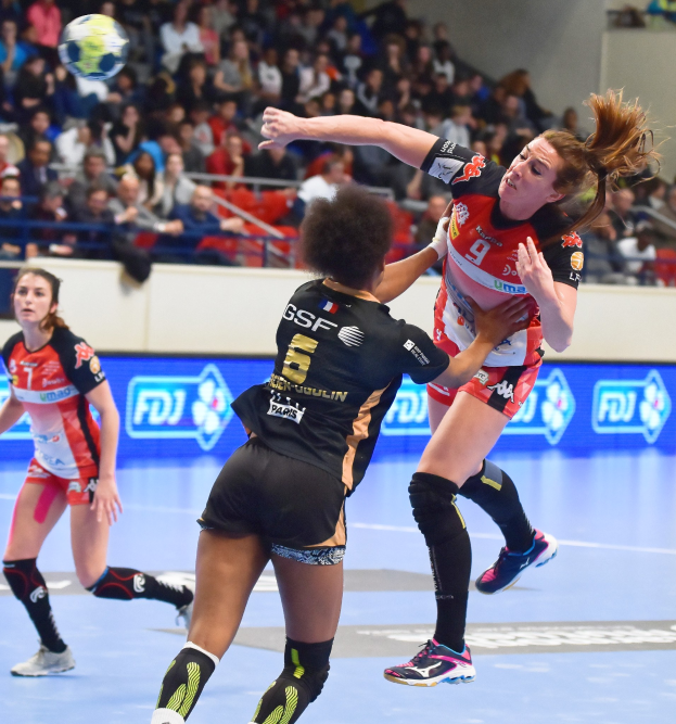 Eine Gruppe von Frauen spielt Handball auf einem Feld mit einem Ball in der Luft, in einer Arena mit Zuschauern im Hintergrund.