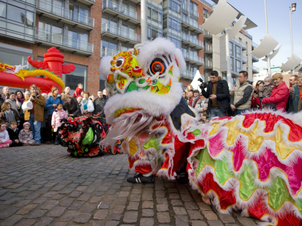 Ein lebendiges chinesisches Neujahrsfest in Amsterdam mit einer Löwentanzvorführung vor einer Zuschauermenge, einige halten Kameras, vor einem Hintergrund aus Gebäuden, Laternenmästen und einem klaren blauen Himmel.