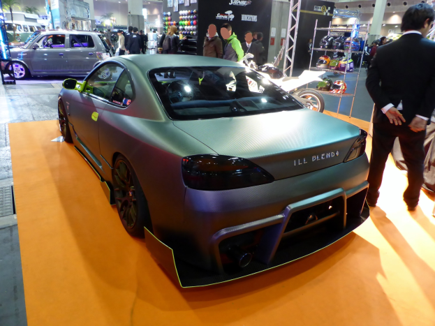 Ein silberner Wagen auf einer Autauschstellung mit Menschen in bunten Kleidern, einem Helmständer und Hintergrundtafeln.