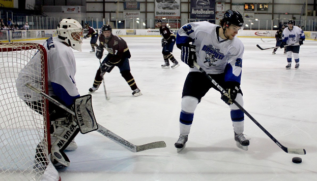 Eine Gruppe junger Männer in Helmen und Eishockey-Ausrüstung, die auf einem Eisplatz mit einem Tor auf der linken Seite spielen, sowie Bannern im Hintergrund und einer Wand.