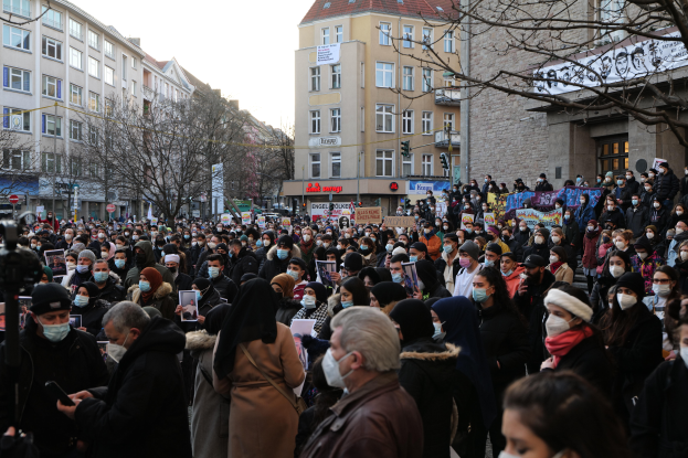 Eine große Gruppe von Menschen mit Mund-Nasen-Schutz steht vor einem Gebäude mit Fenstern, Bäumen, Schildern und Ampeln unter einem bewölkten Himmel, einige halten Schilder und Handys, was auf eine Protestaktion gegen ein Regierungsverbot hinweist.