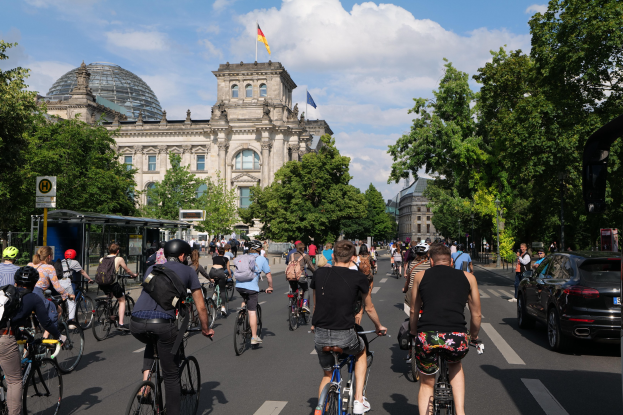 Gruppe von Menschen mit Fahrrädern auf einer von Bäumen gesäumten Straße in Berlin, Deutschland, mit Gebäuden und einer Bushaltestelle, unter einem bewölkten Himmel und einer Flagge auf einem Gebäude.