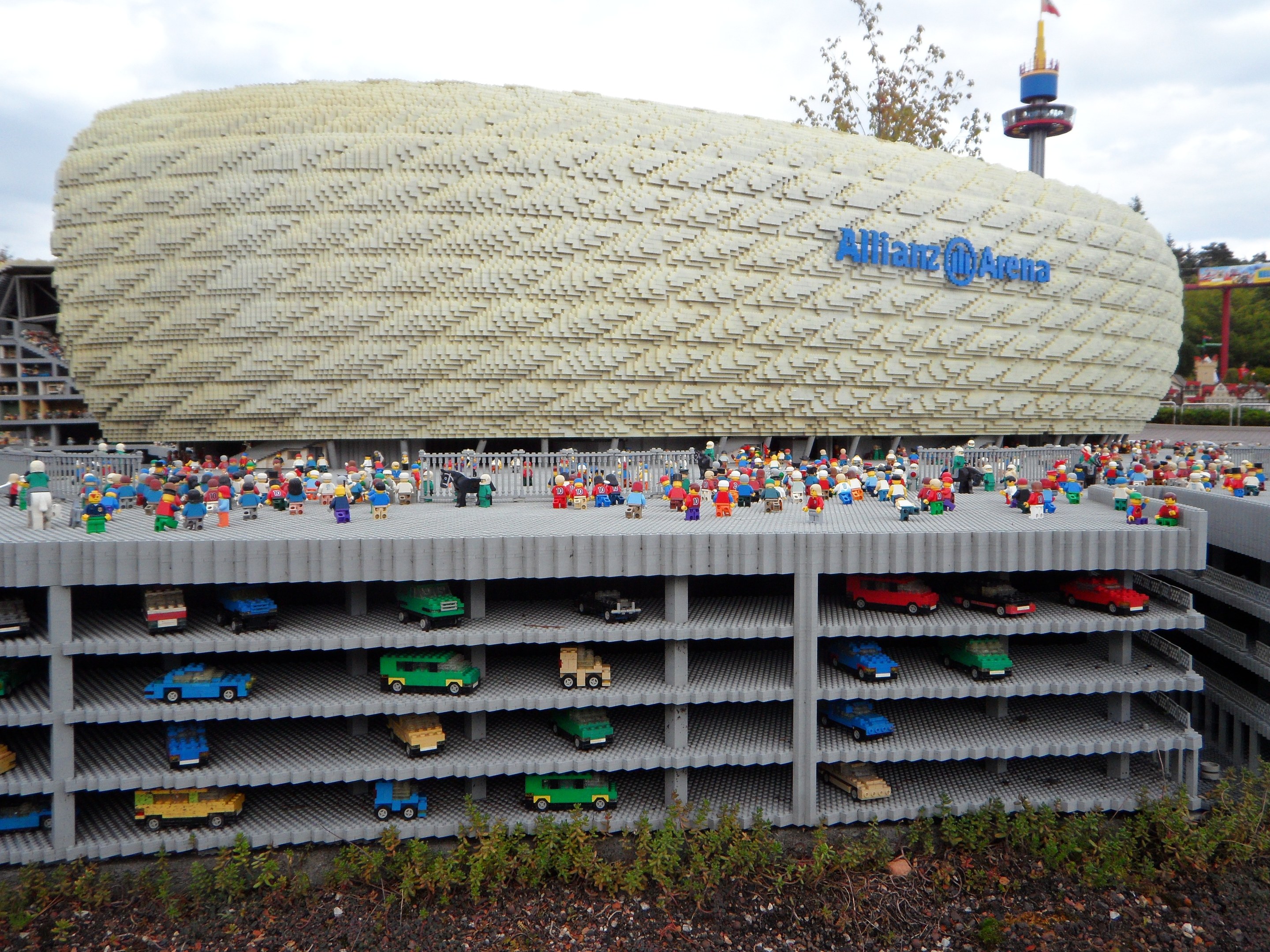 Großes Lego-Modell der Allianz Arena in München, Deutschland, mit Menschen davor, umgeben von Bäumen, Pfählen, einem Turm und Pflanzen, unter einem bewölkten Himmel.