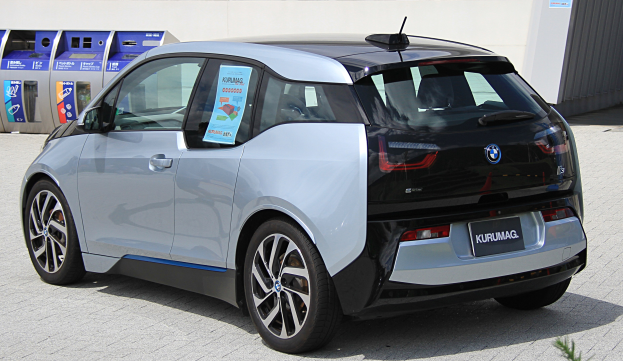 Ein BMW i3 Elektroauto vor einem Gebäude geparkt, mit einer Pflanze im Vordergrund und einer Wand im Hintergrund, das ein Rad in der Mitte des Bildes zeigt.