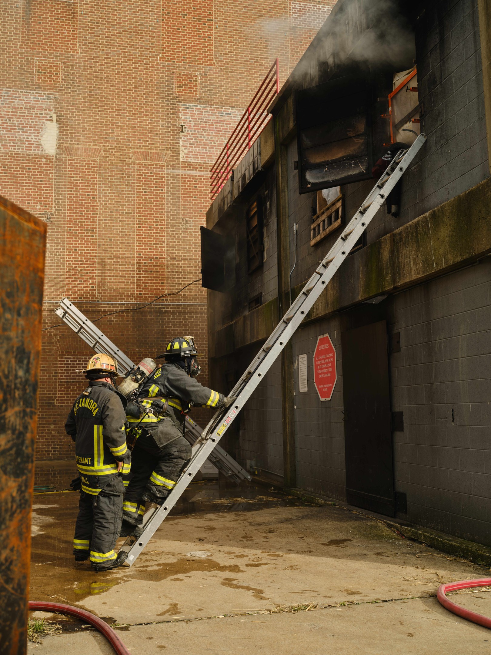Feuerwehrleute mit Helmen klettern an einer Leiter zu einem von Rauch erfüllten Gebäude hoch, mit einem Metallobjekt links und Rohren auf dem Boden.
