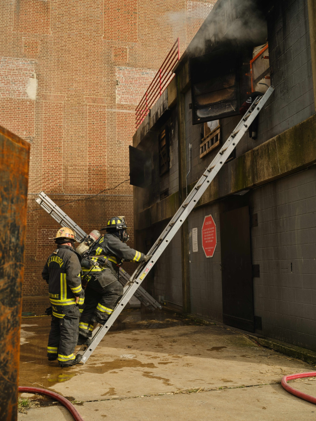 Feuerwehrleute mit Helmen klettern an einer Leiter zu einem von Rauch erfüllten Gebäude hoch, mit einem Metallobjekt links und Rohren auf dem Boden.