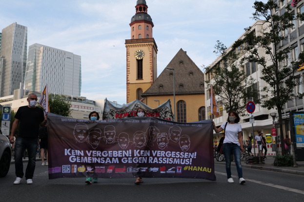 Eine Gruppe von Menschen mit Masken geht eine Straße entlang und hält ein Banner hoch, mit einem geparkten Auto auf der linken Seite, Gebäuden und Bäumen im Hintergrund und einem Kirchturm, der unter einem klaren blauen Himmel sichtbar ist.