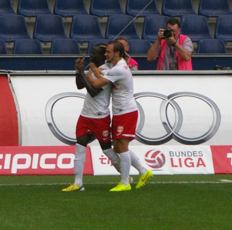Zwei Fußballspieler feiern ein Tor auf einem Feld mit einer Gruppe von Menschen, einem Fotografen, einer Fahne, Stühlen und einer Wand im Hintergrund.