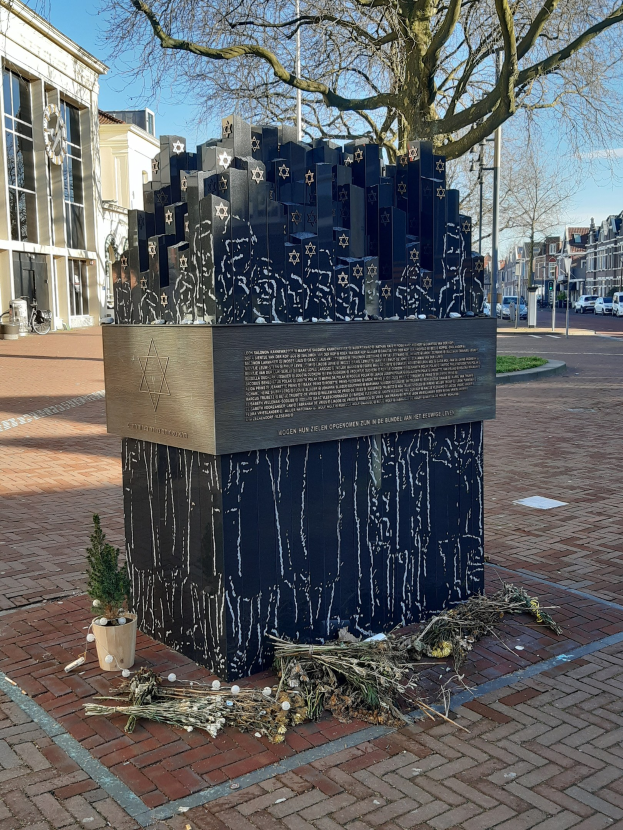 Denkmal für Holocaust-Opfer in einem Stadtplatz mit einem Baum, Topfpflanze, verstreuten Blättern und städtischem Hintergrund einschließlich Gebäuden, Fahrzeugen und Bäumen.