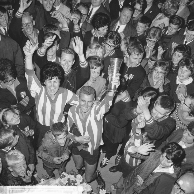 Eine Gruppe von Menschen in Schwarz und Weiß feiert mit einem Pokal, gehalten von der Person in der Mitte, mit Blumen am unteren Bildrand.