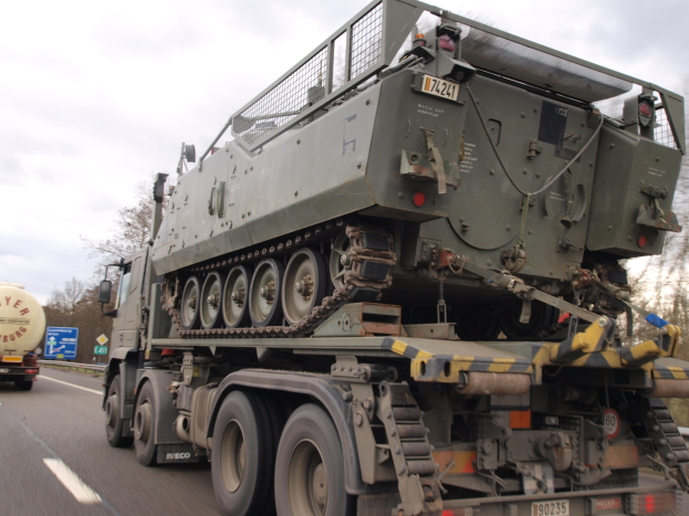 Militärlastwagen transportiert einen Panzer auf seiner Ladefläche, fährt auf einer Autobahn mit Bäumen, Holzplanken und einem klaren blauen Himmel im Hintergrund.