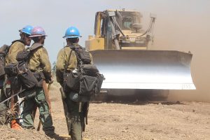 Drei Feuerwehrleute in Helmen und mit Taschen stehen neben einem Bulldozer auf dem Boden, mit Bäumen und einem klaren blauen Himmel im Hintergrund.