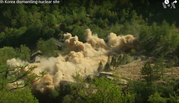Eine große Pilzwolke steigt von einer Kernwaffenexplosion in Nordkorea auf, mit Rauch, der sich über die Landschaft ausbreitet und beschädigte Gebäude und Bäume im Hintergrund sichtbar sind.