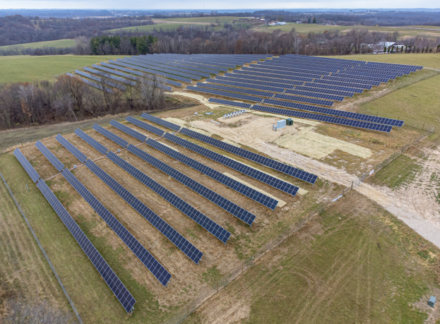 Luftaufnahme eines großen Solarparks im Bau in einem Feld, umgeben von Bäumen und Häusern, mit bewölktem Himmel und Boden voller Solarpanels.