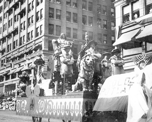 Schwarzes und weißes Foto eines Paradewagen, der mit Statuen von Menschen und Tieren geschmückt ist und die Stra├če einer Stadt entlangfährt, wo Zuschauer von der Seite zusehen.