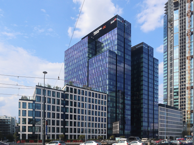 Außenansicht des Deutschen Bank-Hauptsitzes in Frankfurt, Deutschland, mit einem großen mehrfenstrigen Gebäude mit umgebenen städtischen Infrastruktur einschließlich Straßenlaternen, Fahrzeugen und Bäumen unter einem bewölkten Himmel.