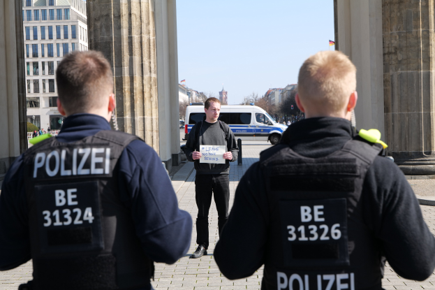 Drei Polizeibeamte stehen vor einem Gebäude, mit einem Mann in der Mitte, der ein Papier hält, flankiert von Säulen und einem belebten städtischen Hintergrund.