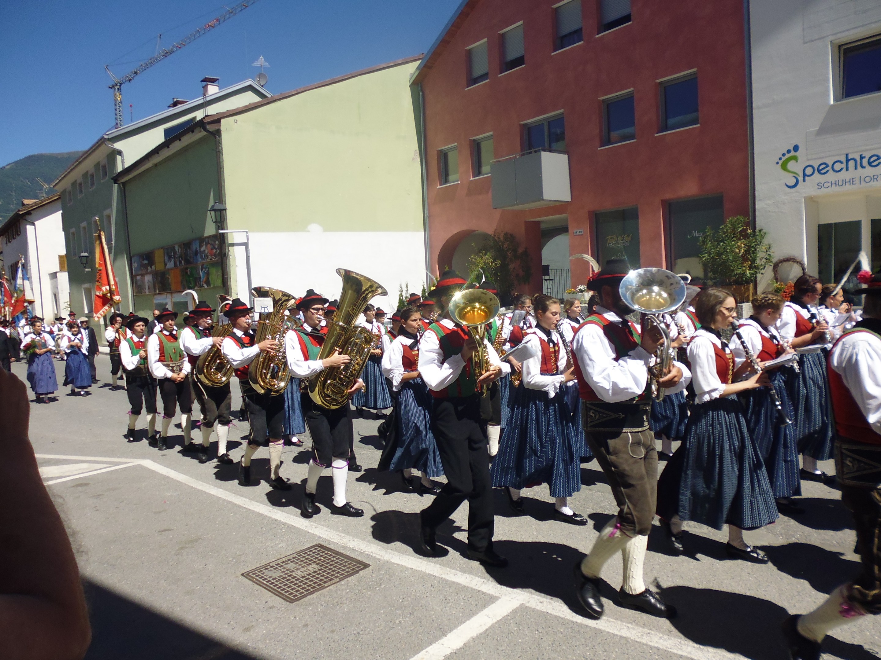Eine Gruppe von Menschen in traditioneller bayrischer Tracht, die auf der Straße mit Musikinstrumenten spielen, während sie durch eine Straße mit Gebäuden gehen, einige halten Fahnen, mit einem Hügel und einem klaren blauen Himmel im Hintergrund.
