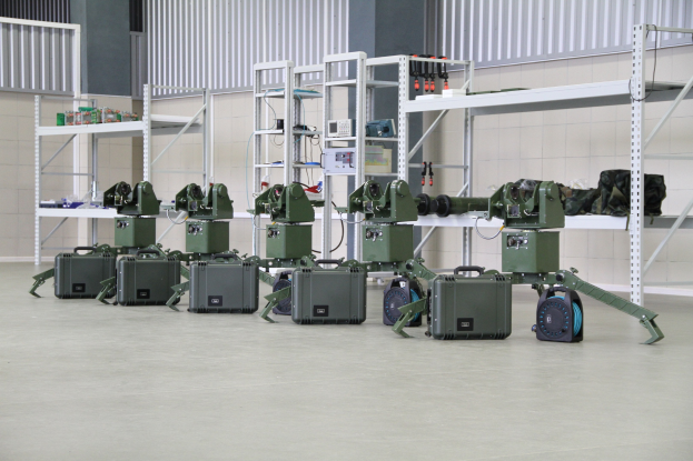 Gruppe von Militärrobotern auf einem Boden mit Koffern und Objekten auf Gestellen, positioniert in der Nähe einer Wand und eines Geländers als Teil eines Verteidigungssystems.